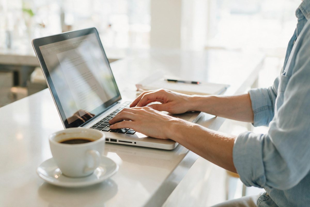 Owner typing on a laptop at a bright counter with a coffee, drafting text